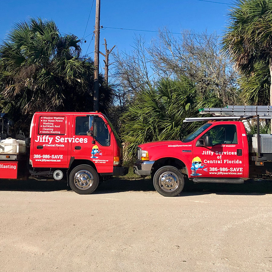Two red service trucks for Jiffy Services Central Florida are parked side by side on a dirt lot, with palm trees and clear blue sky in the background. The trucks display company information and a cartoon handyman logo.